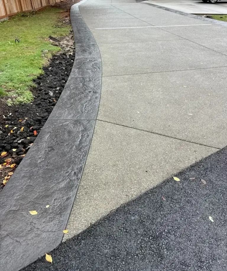 Curving charcoal stamped concrete border separating the exposed aggregate driveway from the lawn.