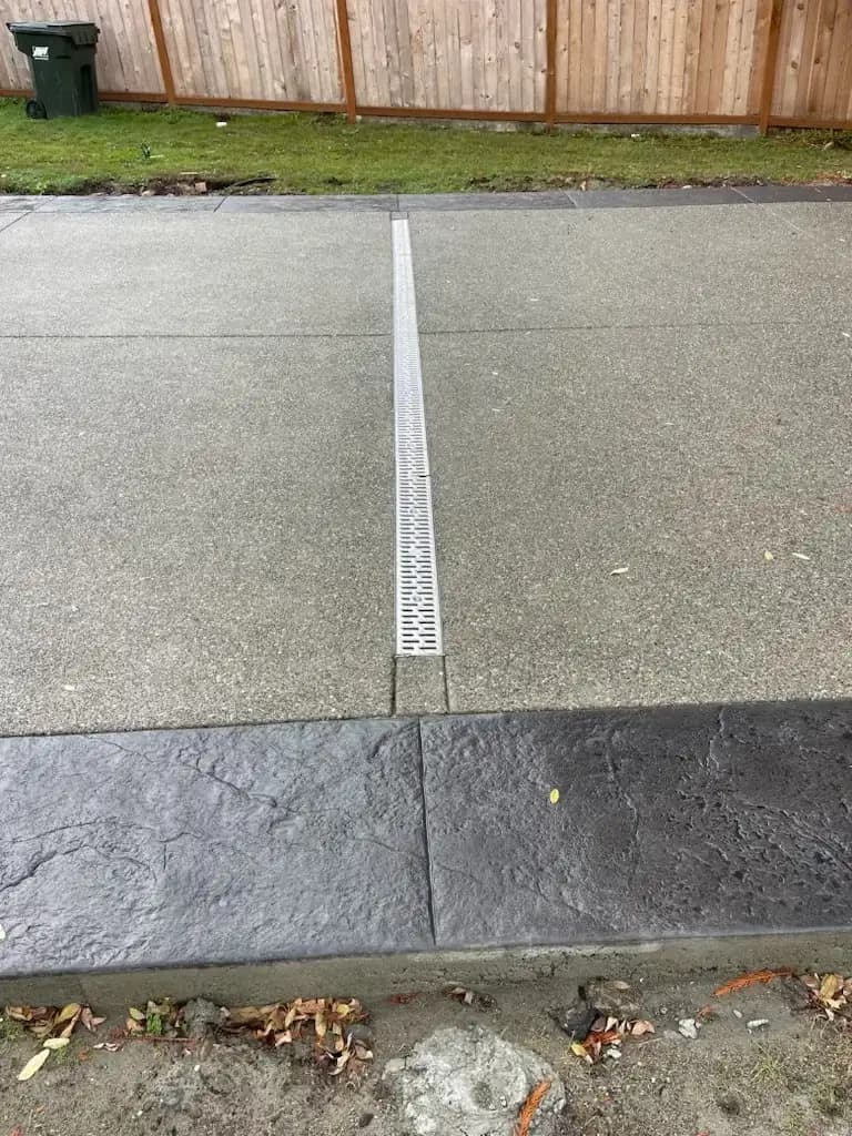 Close-up of a metal channel drain set into the exposed aggregate concrete driveway surface.