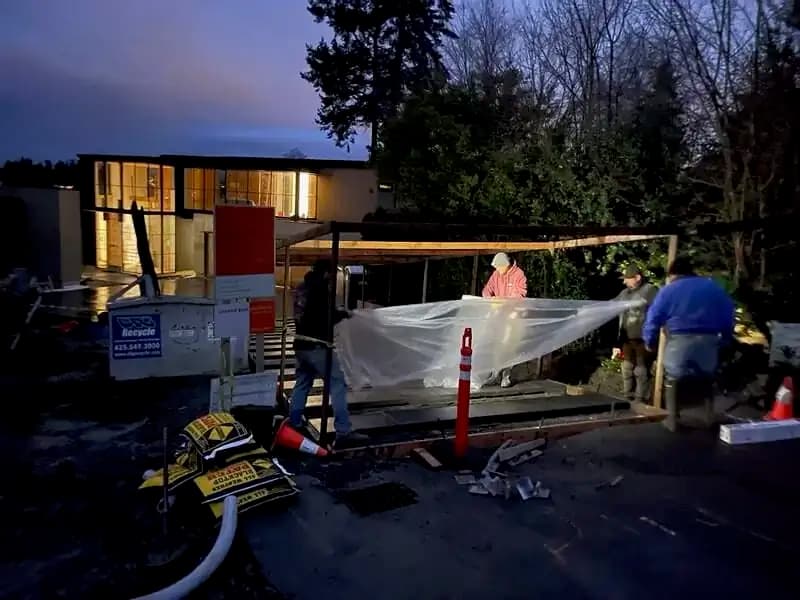 Construction crew covering a section of newly poured concrete at dusk under a temporary wooden frame.