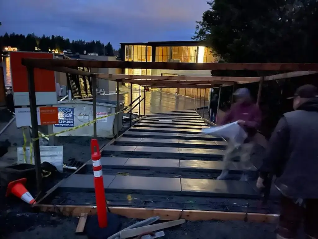 A crew pulling a plastic sheet over a freshly poured and formed concrete walkway during the evening.