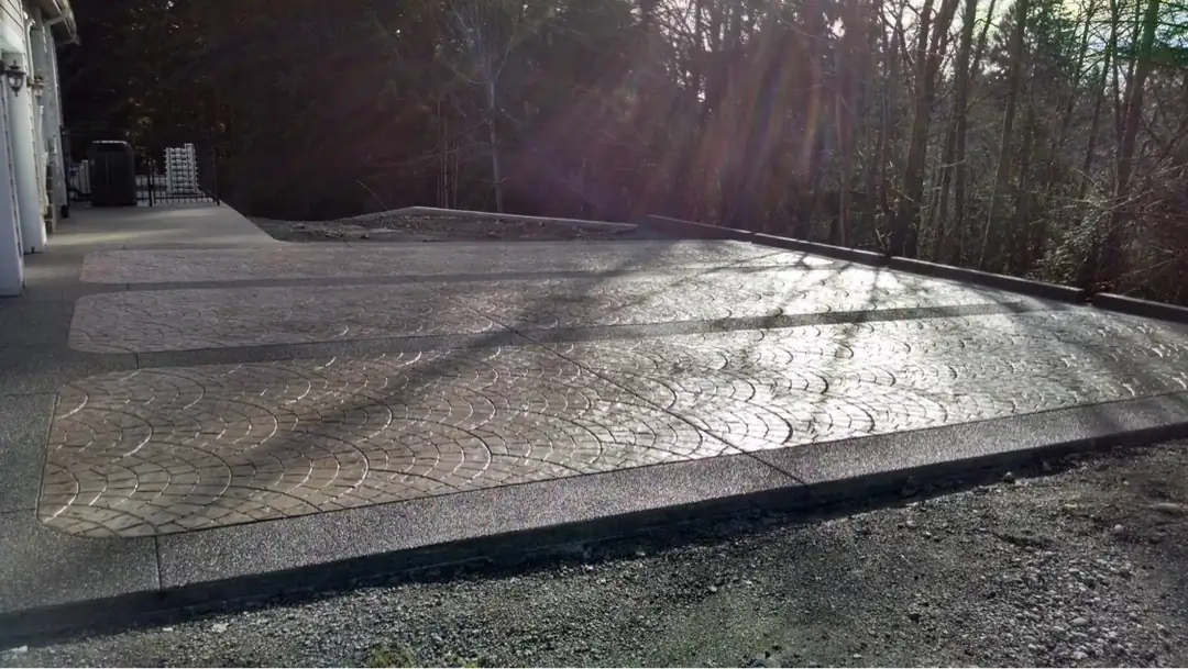 Sunlight reflects off the sealed surface of a new stamped and bordered concrete driveway next to a wooded area.