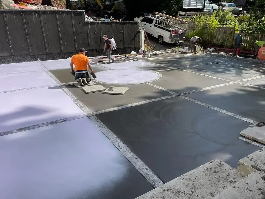 Workers applying a finishing spray to the freshly poured concrete pads surrounding the central inlay.