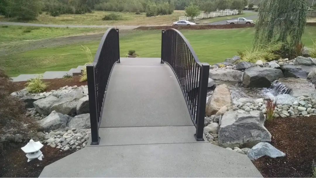 An arched concrete pedestrian bridge with metal railings spanning a rock-lined stream in Puyallup, WA.