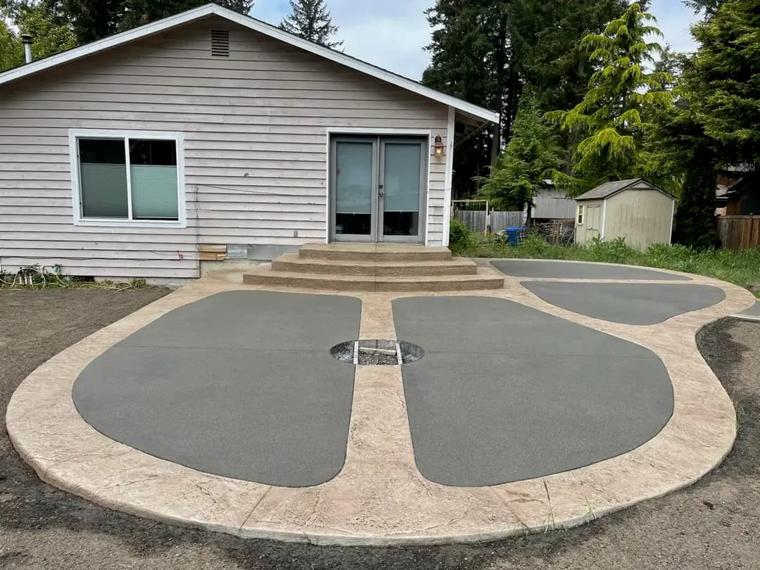 Wide view of a residential backyard featuring a new custom-shaped concrete patio with a two-tone design.