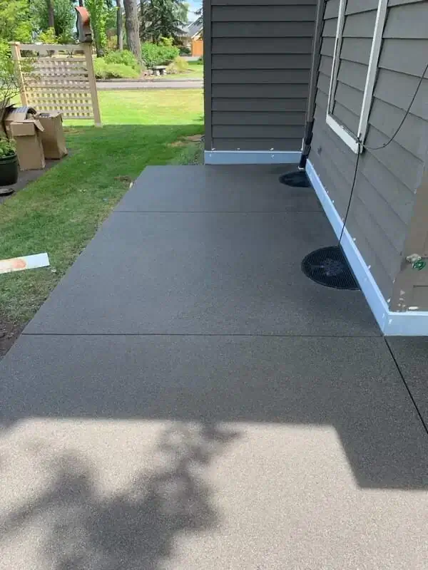 A narrow concrete walkway with a consistent sand finish running alongside a house with gray siding.