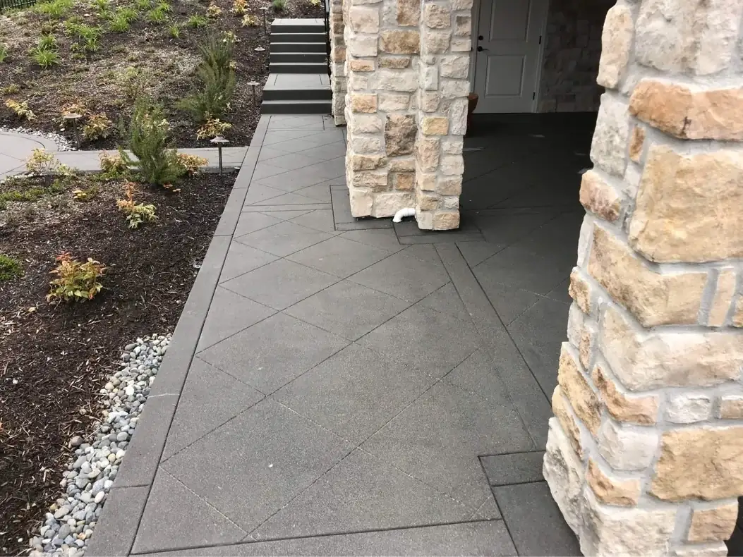 Exposed aggregate walkway with a diamond saw-cut pattern running alongside stone pillars of a covered porch.